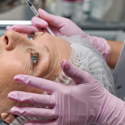 Close-up of a senior woman receiving a cosmetic injection in a clinical setting with gloves and a hairnet.